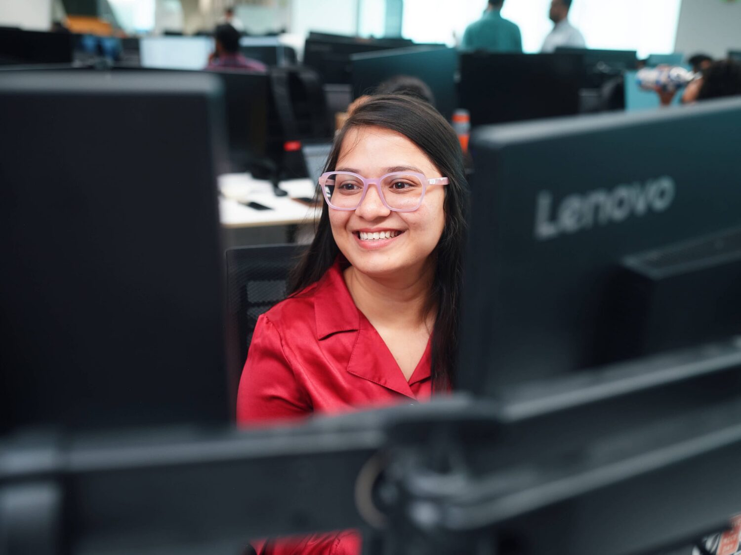 A smiling woman in a red shirt sits at a computer workstation in a lab with Lenovo monitors.