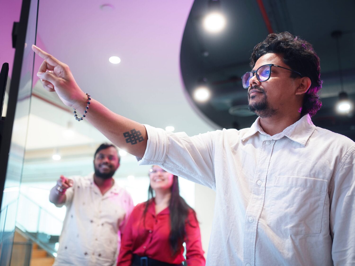 A man in a white shirt and glasses points to the left in a modern indoor space, with two people standing in the background.