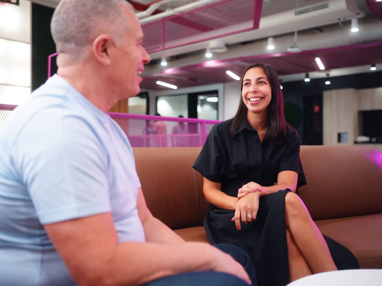 A man and a woman sit on a brown sofa in a modern indoor lounge, smiling and chatting.
