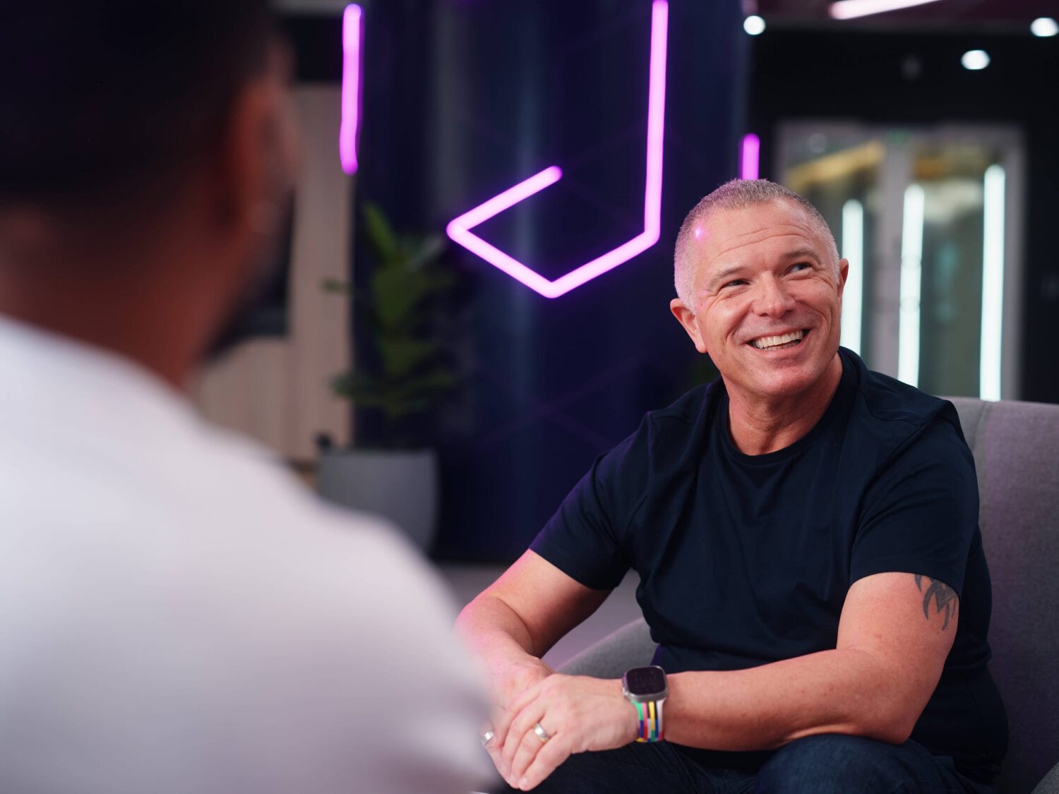 Smiling man in a black T-shirt sits and chats with another person in a modern office setting with purple neon lights.