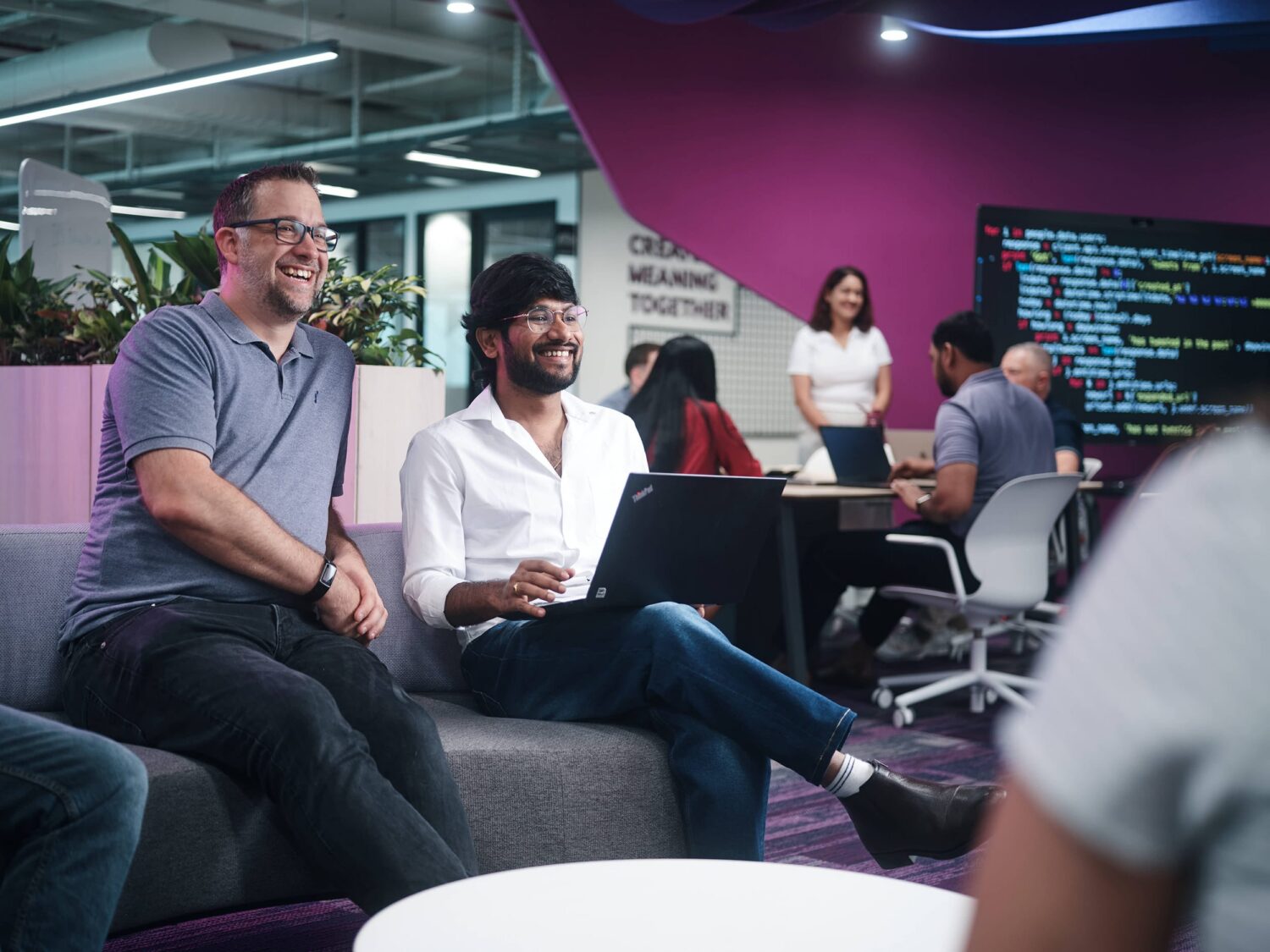 Two men sit on a gray sofa with laptops in a bright, modern office as other colleagues work near a purple accent wall.