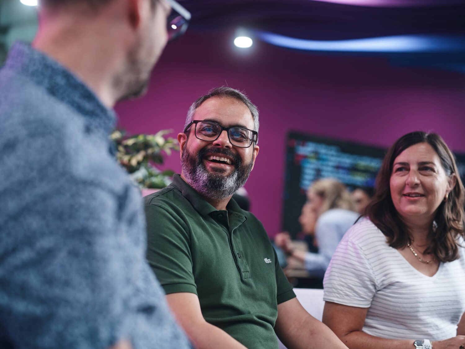 Three adults sit together and smile in a colorful indoor setting, with a bearded man in a green polo and a woman in a white striped shirt.
