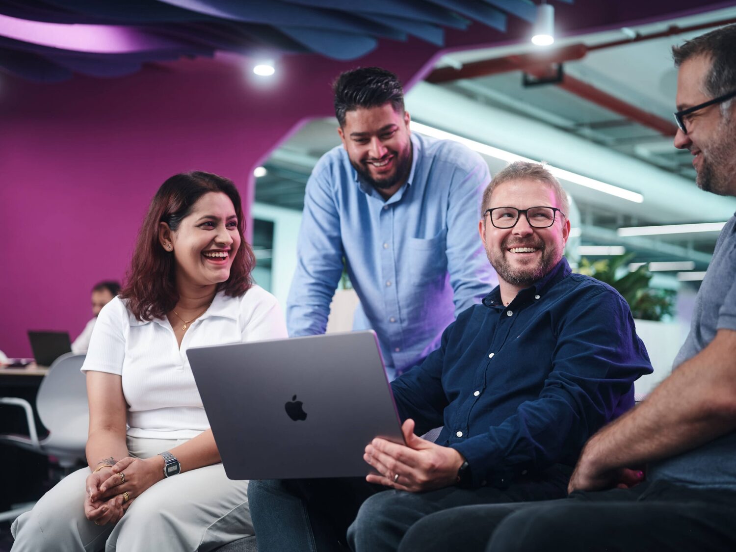 Diverse group of coworkers gathered around a laptop, smiling during a collaborative office meeting.