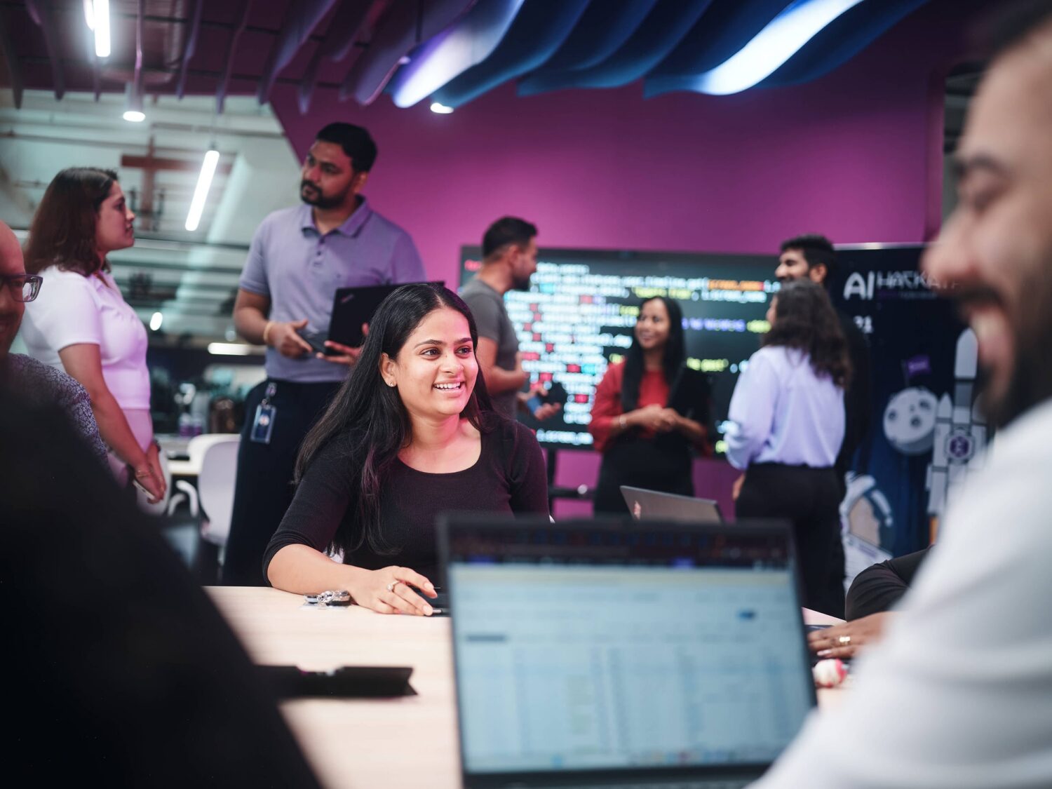 A woman smiles at a laptop on a table in a busy, colorful modern office with coworkers chatting in the background.