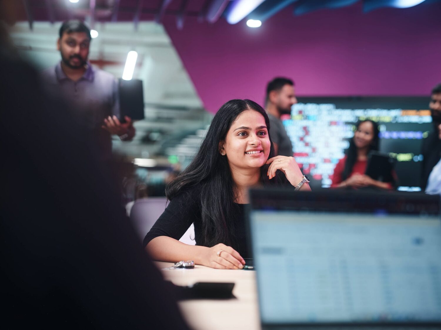 A smiling woman with long dark hair sits at a desk in a busy office, with colleagues and computer screens visible in the background.