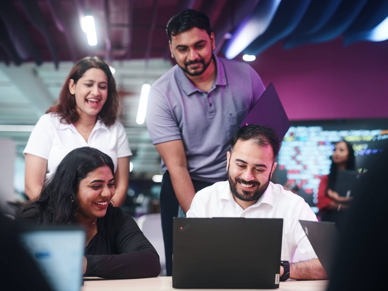 Four coworkers gather around a laptop in a modern, colorful office, smiling as they collaborate.