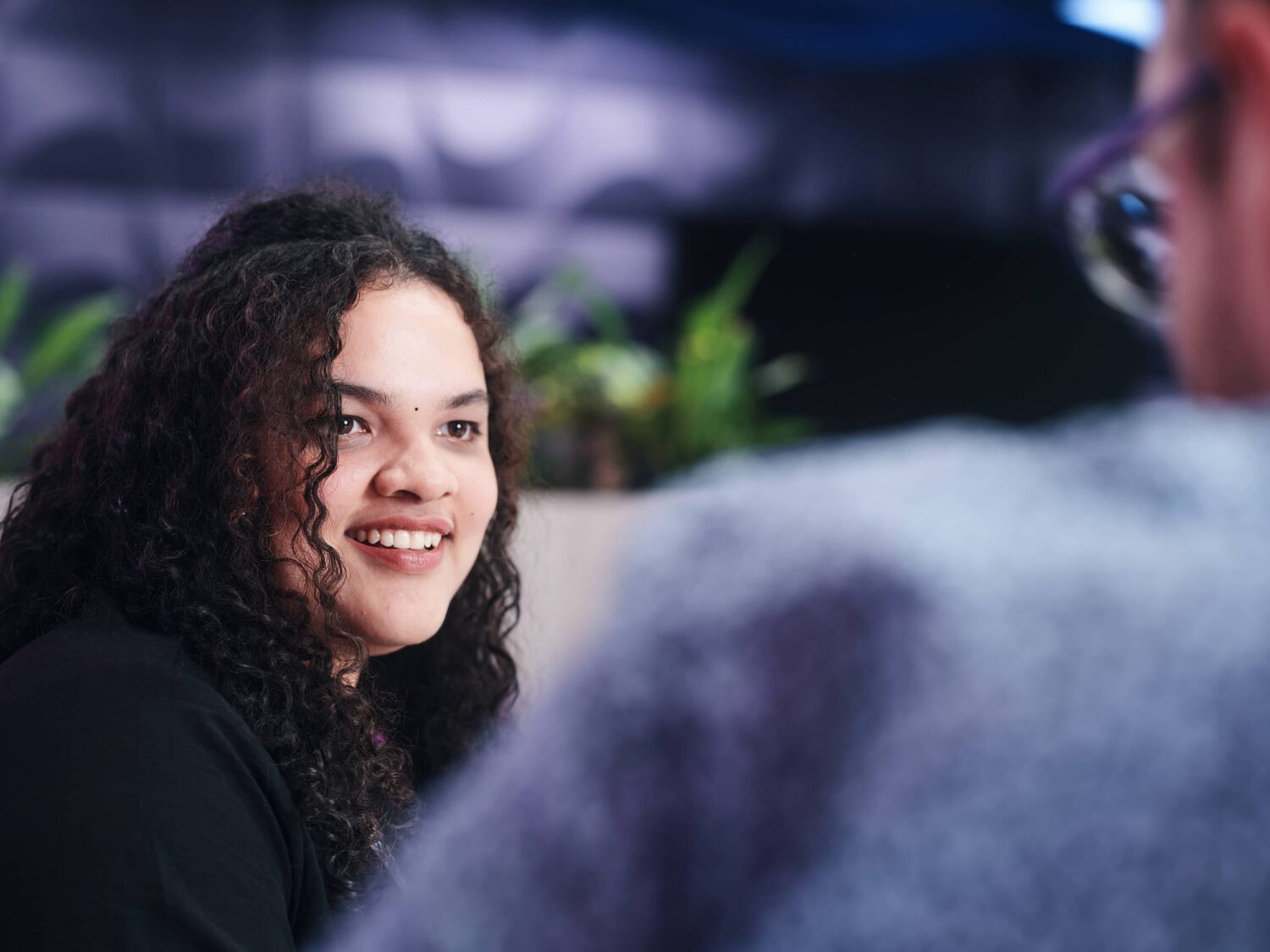 A smiling young woman with curly hair talks to a person whose back is to the camera in an indoor setting.
