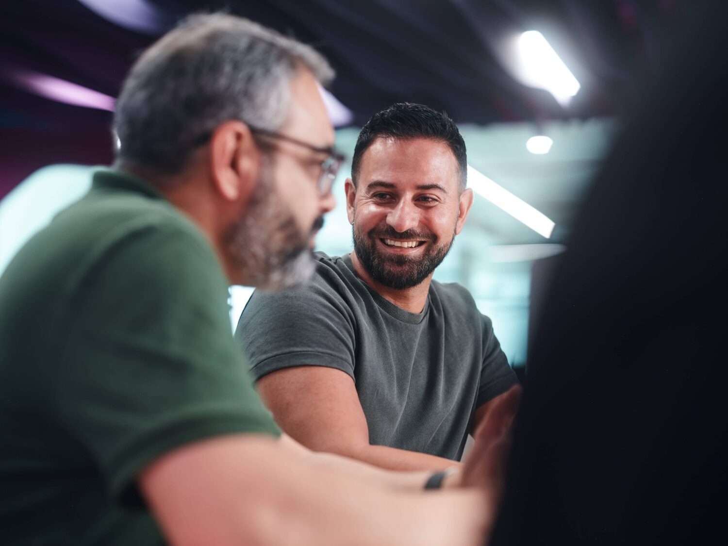 Two men sit at a table in a modern office, smiling and chatting.