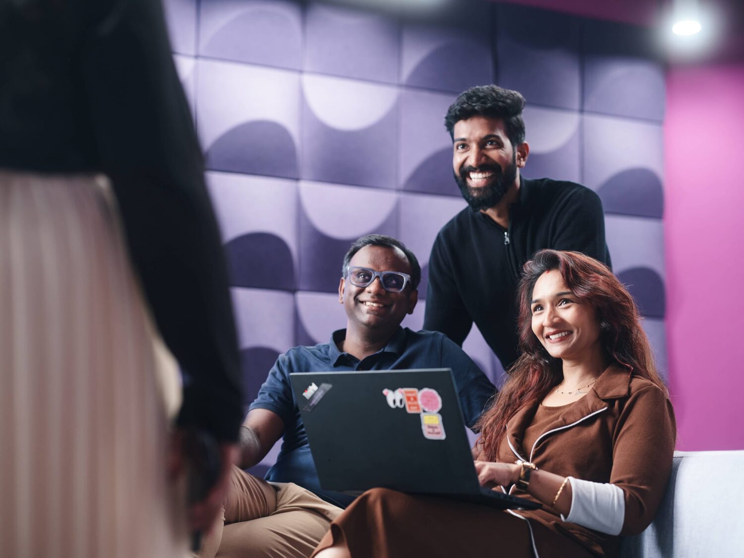 Three coworkers smile at a laptop in a modern studio with a purple geometric wall.
