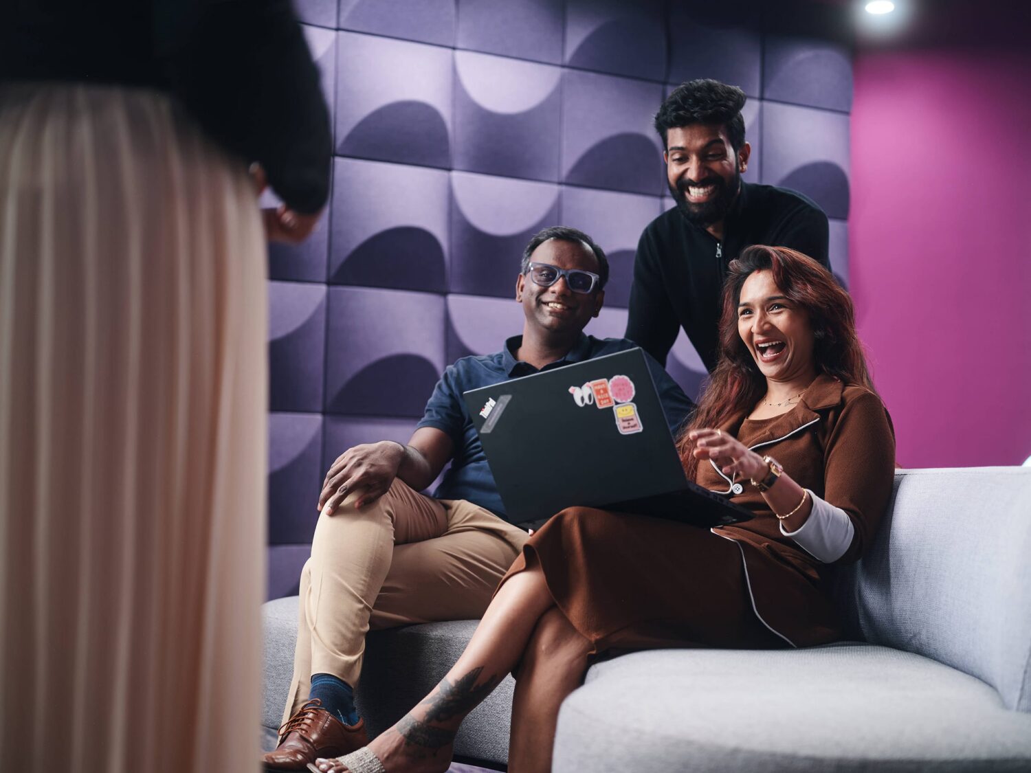 Three people sit on a white sofa in a modern room, gathered around a laptop.