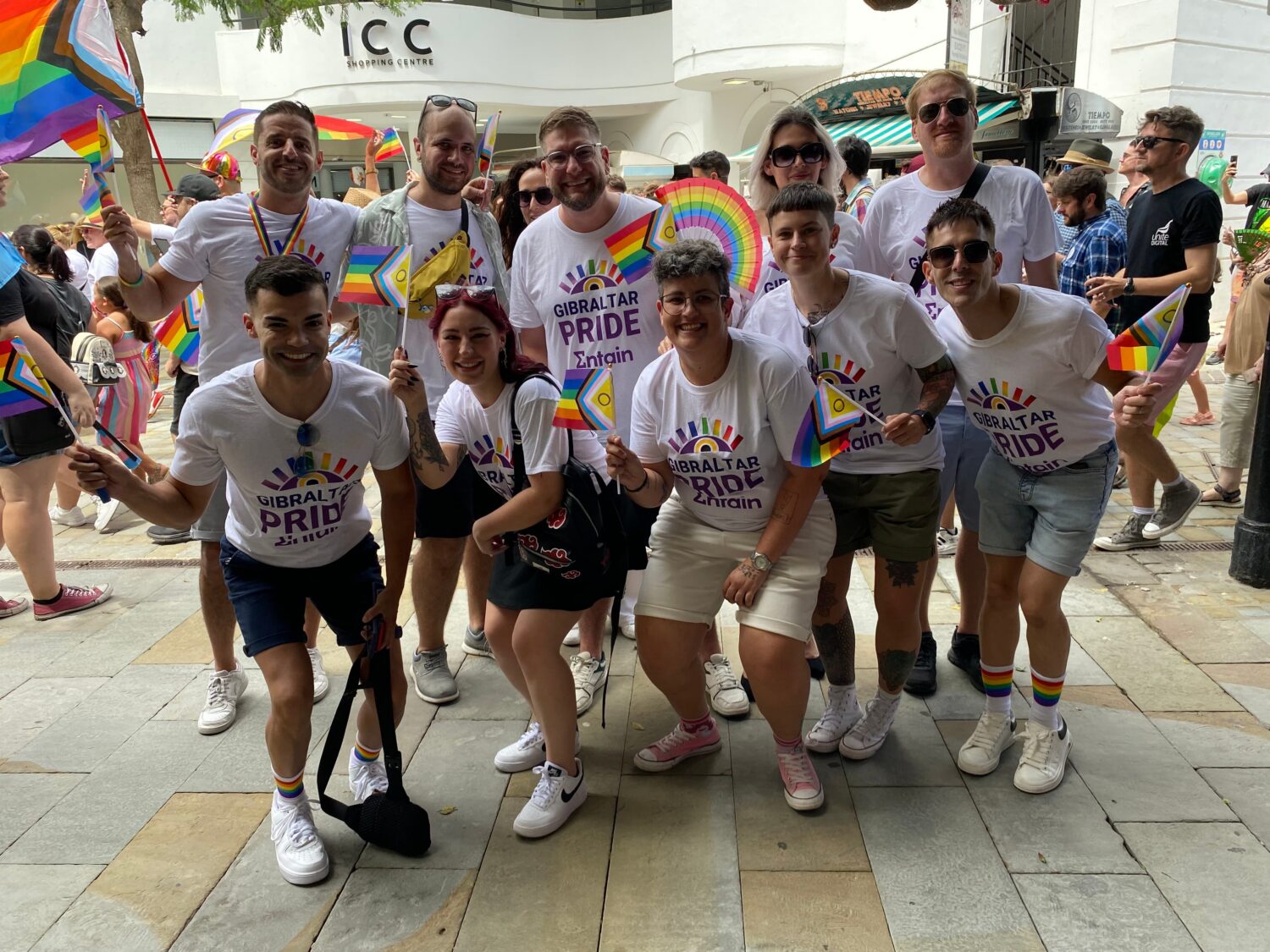 Group of diverse people wearing white Pride shirts pose together on a sunny sidewalk at a Pride event, holding rainbow flags.