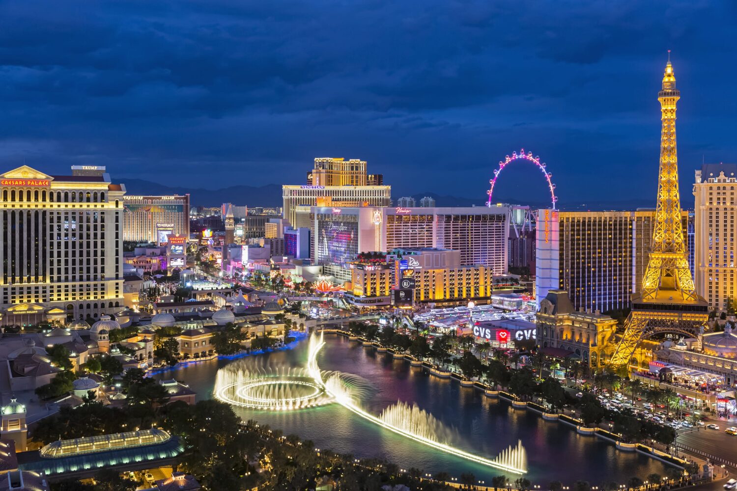 Nighttime view of the Las Vegas Strip with the Bellagio fountains in the foreground, the Eiffel Tower replica at Paris Las Vegas, and the High Roller observation wheel.