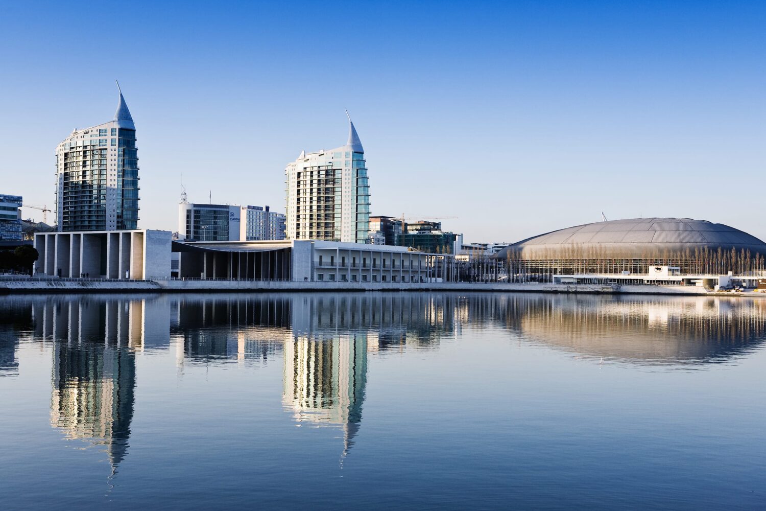 Modern city skyline along a waterfront with glass skyscrapers reflected in calm water.
