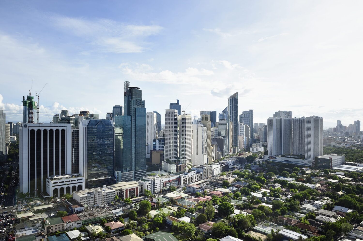 A dense urban skyline with numerous tall skyscrapers and a foreground of green trees and low-rise houses under a bright blue sky.