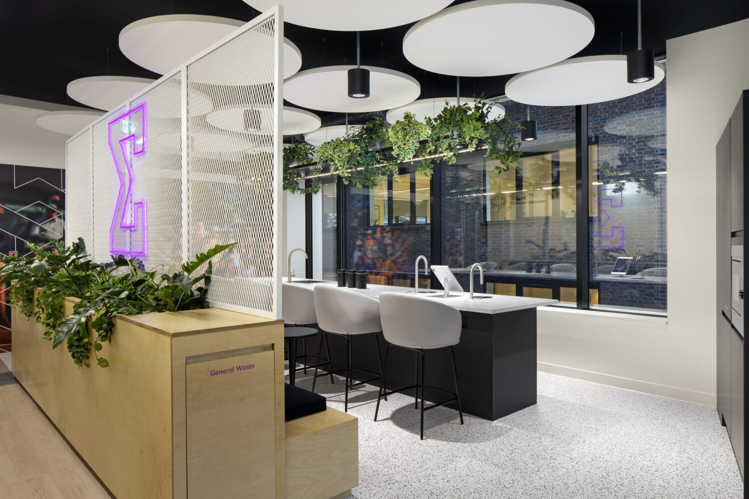 Modern office lounge featuring white circular ceiling panels, greenery, and a row of white chairs at a long table beside large windows.
