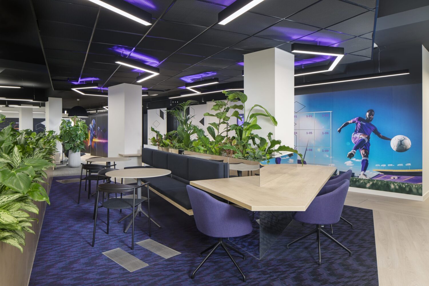 A modern coworking space with purple chairs, wooden tables, lush plants, neon purple ceiling lights, and a blue mural of a person with a soccer ball.