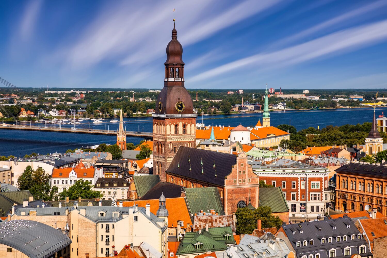 European cityscape with a tall brick church tower, red-tiled roofs, and a river with a bridge.