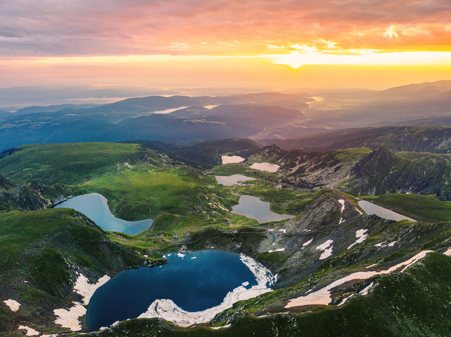 Alpine landscape at sunset with green valleys, snow patches, and multiple blue crater lakes nestled in the mountains.