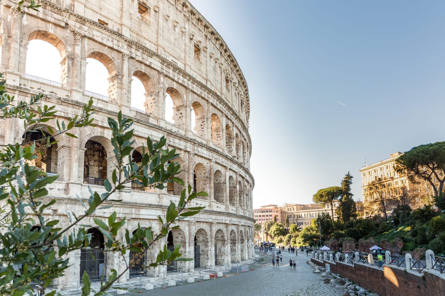 Ancient Colosseum on the left with a tree-lined street and people walking on the right under a clear blue sky.