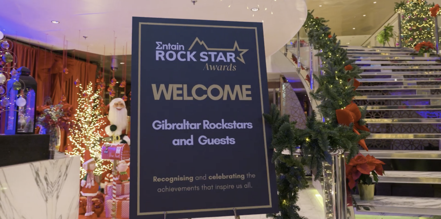 Blue welcome sign in a festive indoor venue reads "Welcome Gibraltar Rockstars and Guests" with holiday decorations and a stairway to the right.