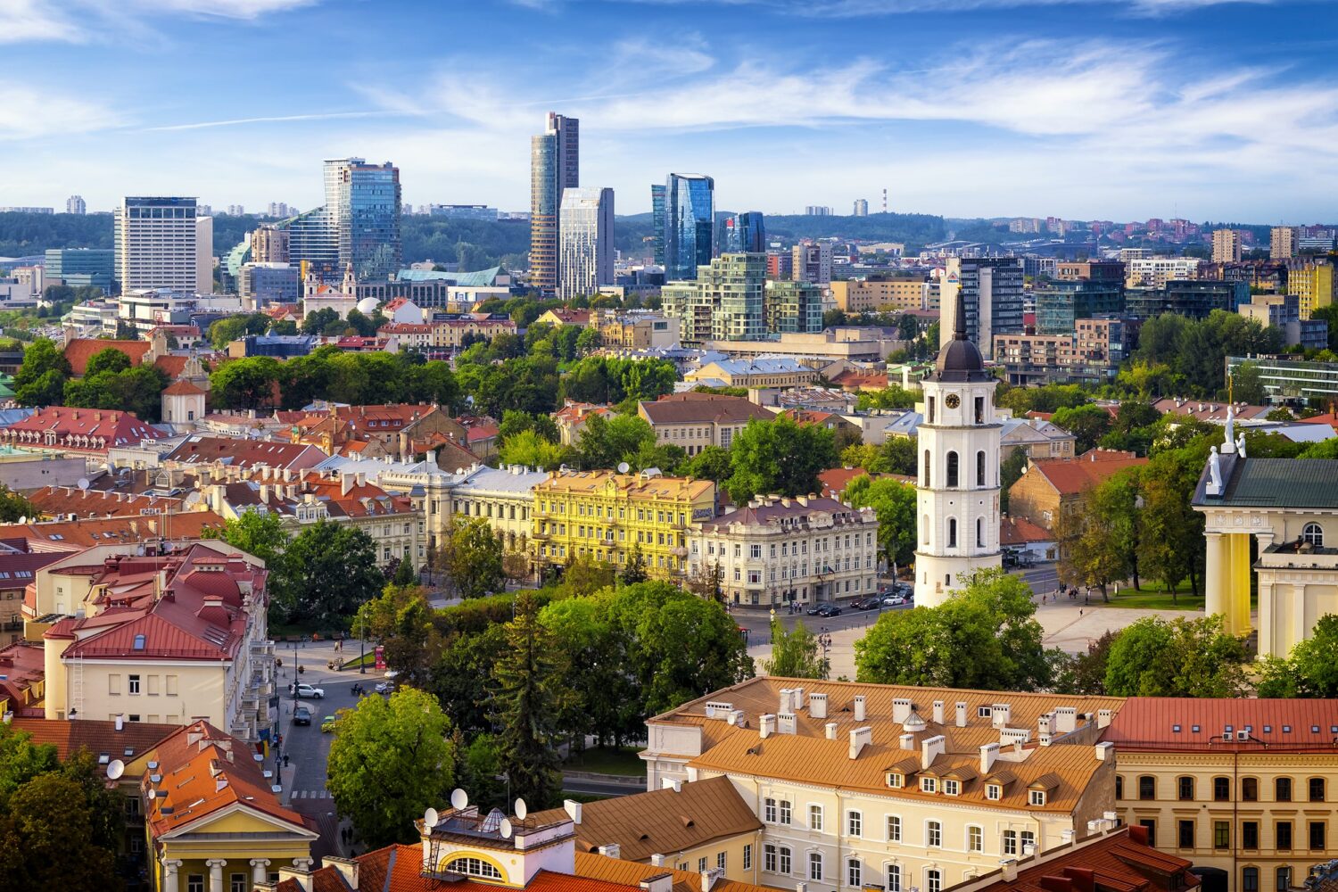 Panoramic cityscape with red-tiled rooftops, a prominent white church tower, green trees, and a blue sky.
