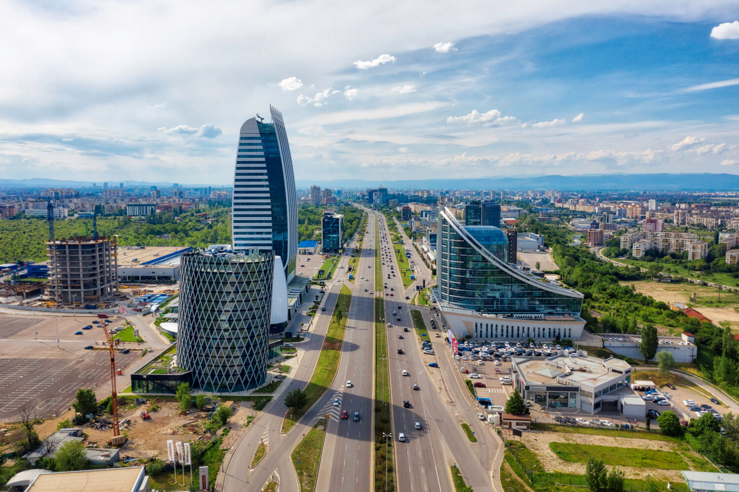 Aerial view of a city with a wide, divided highway flanked by modern high-rise buildings and green parks under a blue sky.